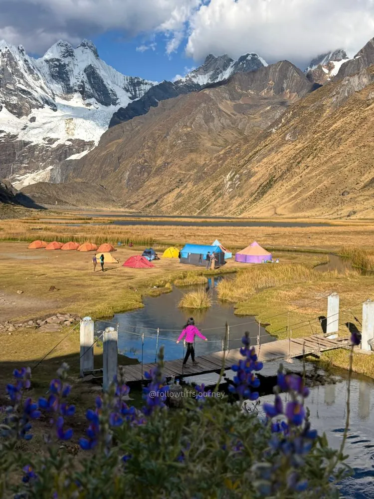 Tiff crossing a small bridge toward colourful trekking tents in an alpine valley