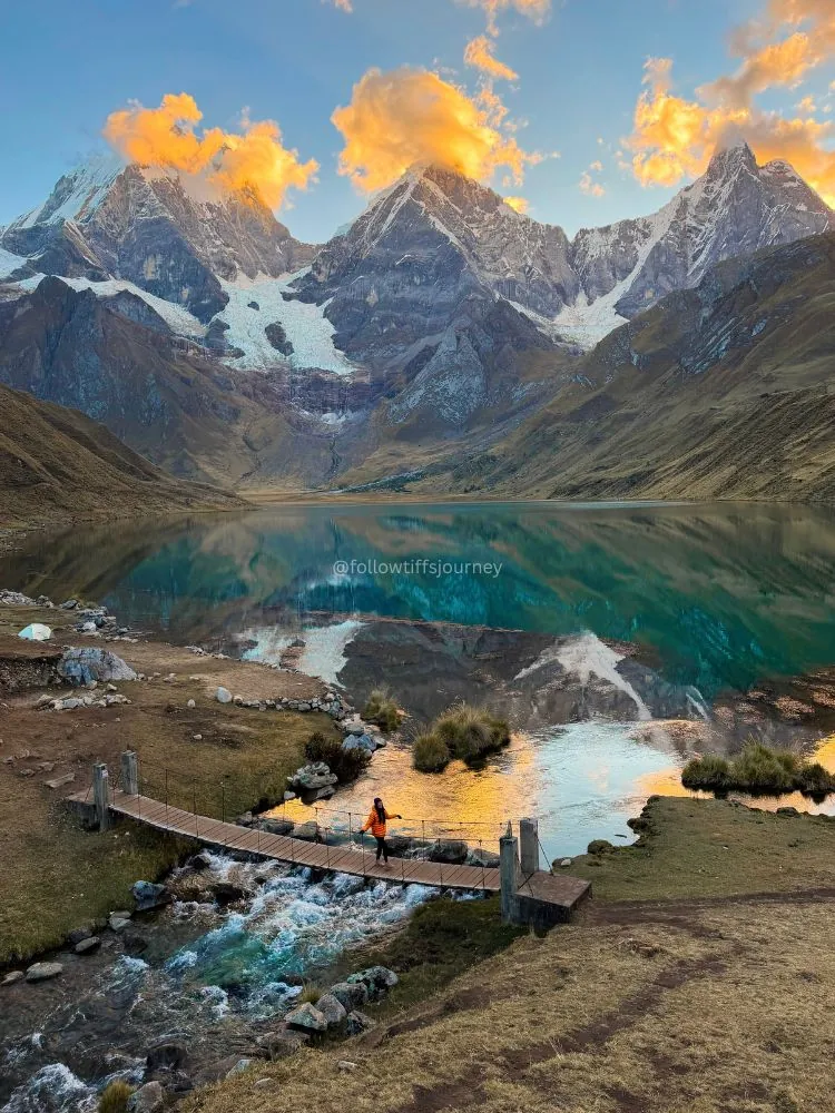 Sunset light on Huayhuash peaks reflected in a lake, with a hiker on the foreground bridge